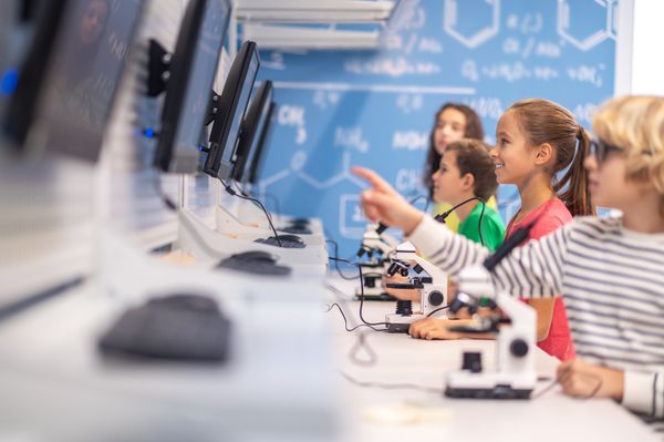 Children learning in a classroom, by using microscopes to look at cell samples and record their interactions over the computer.