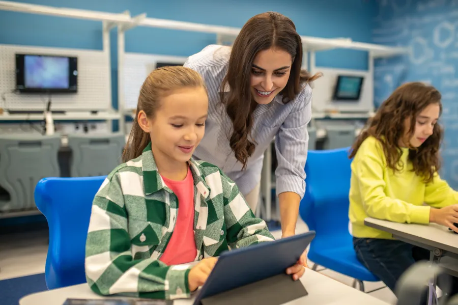 A kind teacher helping a happy young student over a tablet.