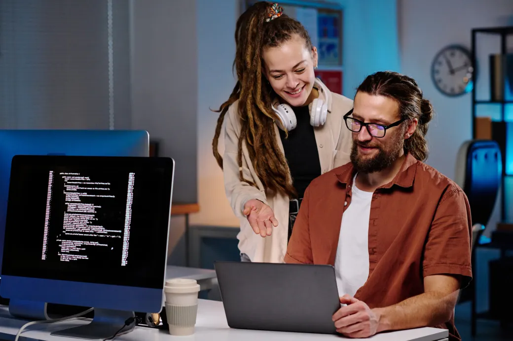 An older, and younger individual both looking confidently collaborating at a laptop, while in an office space.