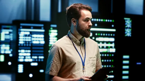 Employee with an ID badge, in a data center with a tablet, monitoring technical infrastructure.