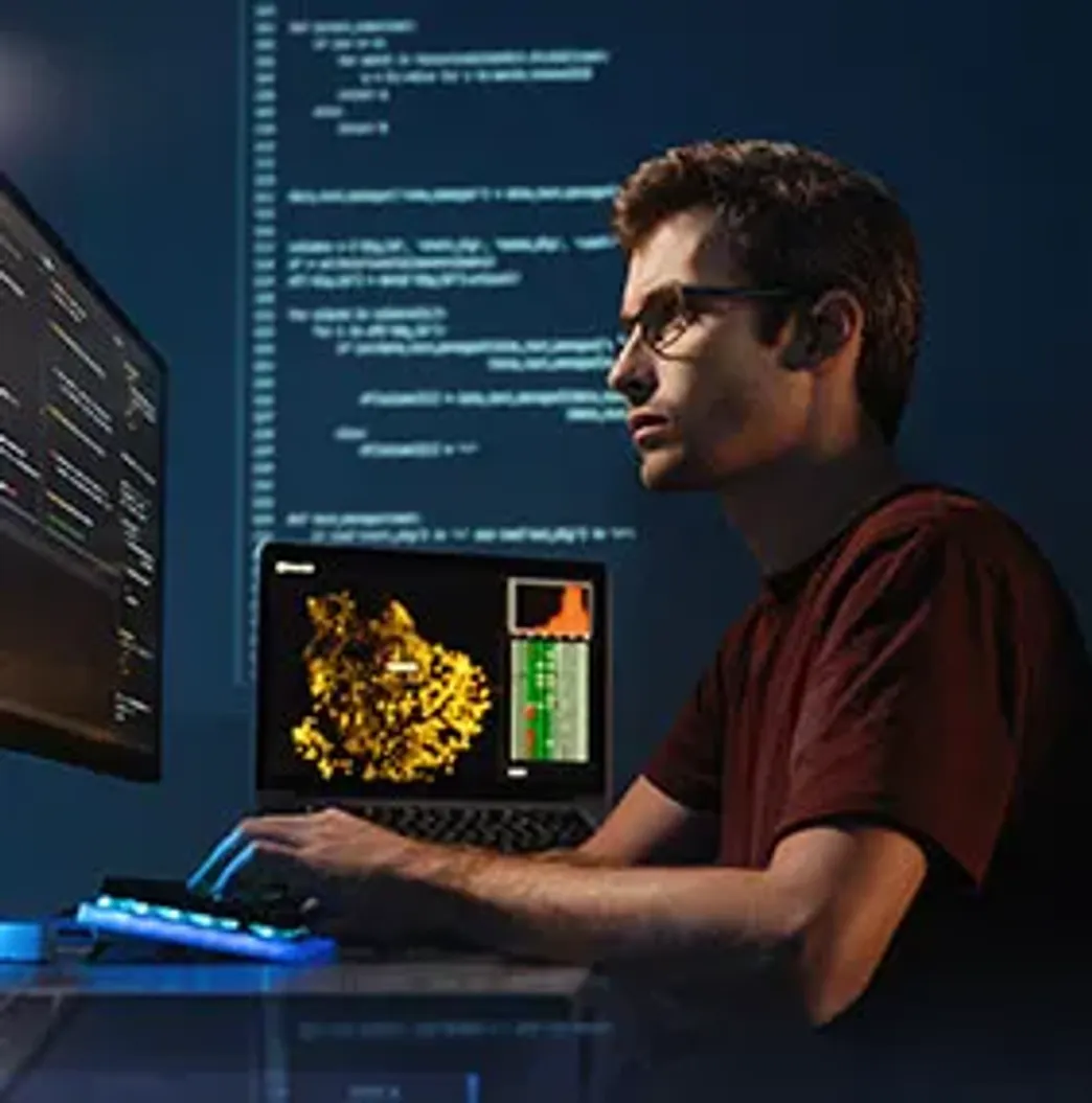 Person in a dark environment working at a computer setup. A display of data is on another monitor and the atmosphere is focused.
